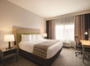 Bright hotel room with a large bed, striped runner, bedside tables with lamps, a desk with chair, and a window with sheer curtains. Neutral decor creates a welcoming atmosphere.