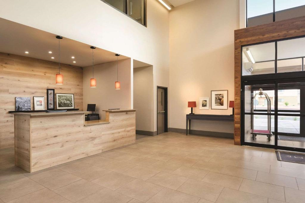 Hotel lobby with a wooden reception desk, pendant lights, and tiled flooring. Glass entrance on the right, framed artworks, and a luggage cart are visible.