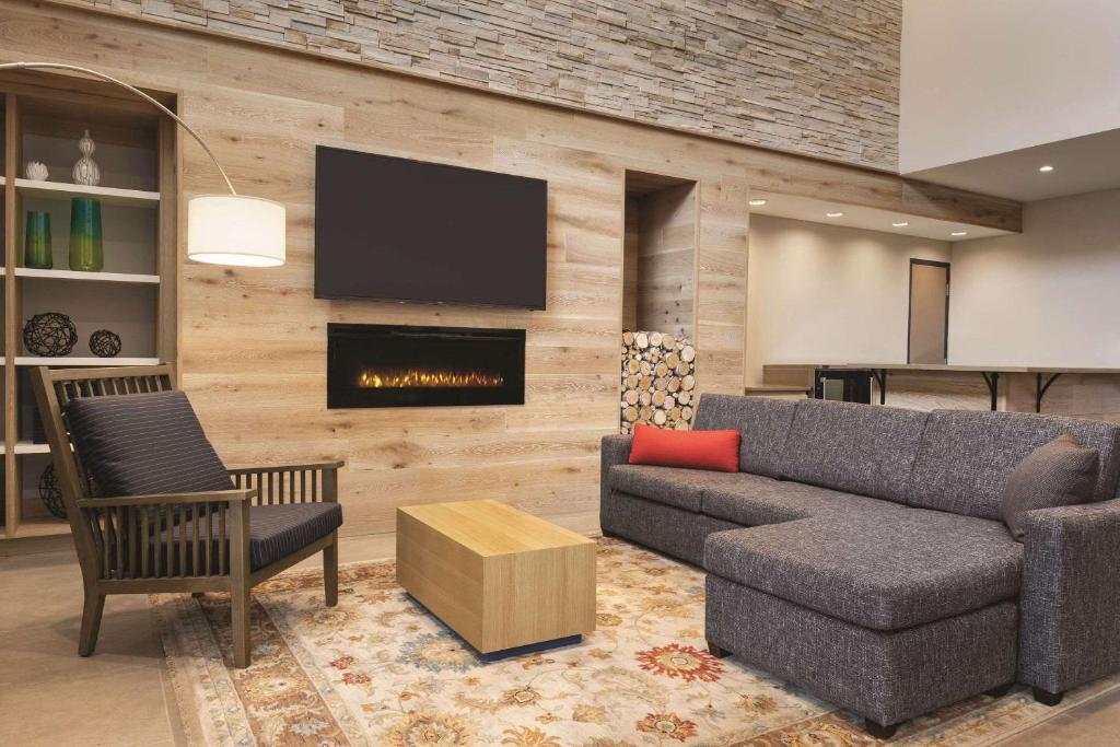 A modern living room with a gray sectional sofa, wooden armchair, and rectangular coffee table. A flat-screen TV and electric fireplace are mounted on a wood-paneled wall.