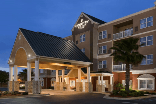Hotel entrance with a covered driveway, illuminated at dusk. Four-story building with balconies and a palm tree nearby. The sign on the building reads "Country.