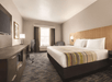 Hotel room with a king-sized bed, striped runner, TV, desk with a chair, armchair, and window with curtains. Neutral tones and modern decor.