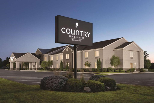 Exterior view of a Country Inn & Suites by Radisson at dusk, featuring a well-lit sign and an empty parking lot, surrounded by neatly maintained shrubs and lawn.