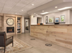 Hotel lobby with a wooden reception desk, clock, and fireplace. Decorative photos and seating area on the left. Markings on the floor for social distancing.