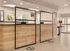 Hotel reception area with wooden counter, transparent partitions, and social distancing floor markers; artwork on walls and a red lamp in the background.