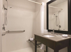 A hotel bathroom with a walk-in shower, a mounted showerhead, a safety grab bar, a towel on a shelf, a sink with toiletries, and a large mirror.