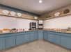 Breakfast bar with a drink dispenser, fruits, pastries, and yogurt on blue counters and tiled walls. Plates and decor are displayed on shelves above.