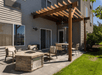 A concrete patio with a fire pit with chairs around it and a table and chairs with a small awning over it on a sunny day in Portland.