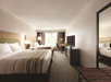Hotel room with two double beds, a TV on a dresser, a desk with a chair, and a large mirror. A window with curtains provides natural light.