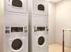 Two stacked white industrial washing machines and dryers in a laundry room with beige walls and tile flooring.