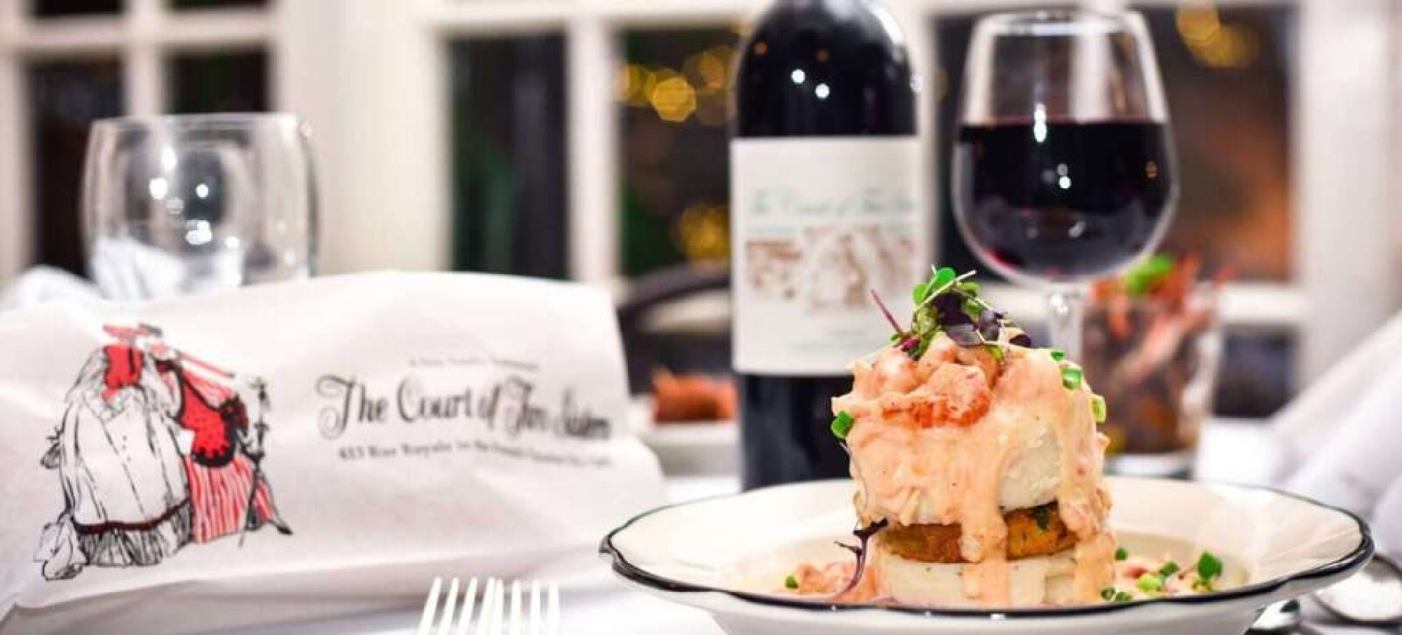 A plated seafood dish with sauce, a glass and bottle of red wine, and a napkin displaying "The Court of Two Sisters" restaurant logo on a formal dining table.