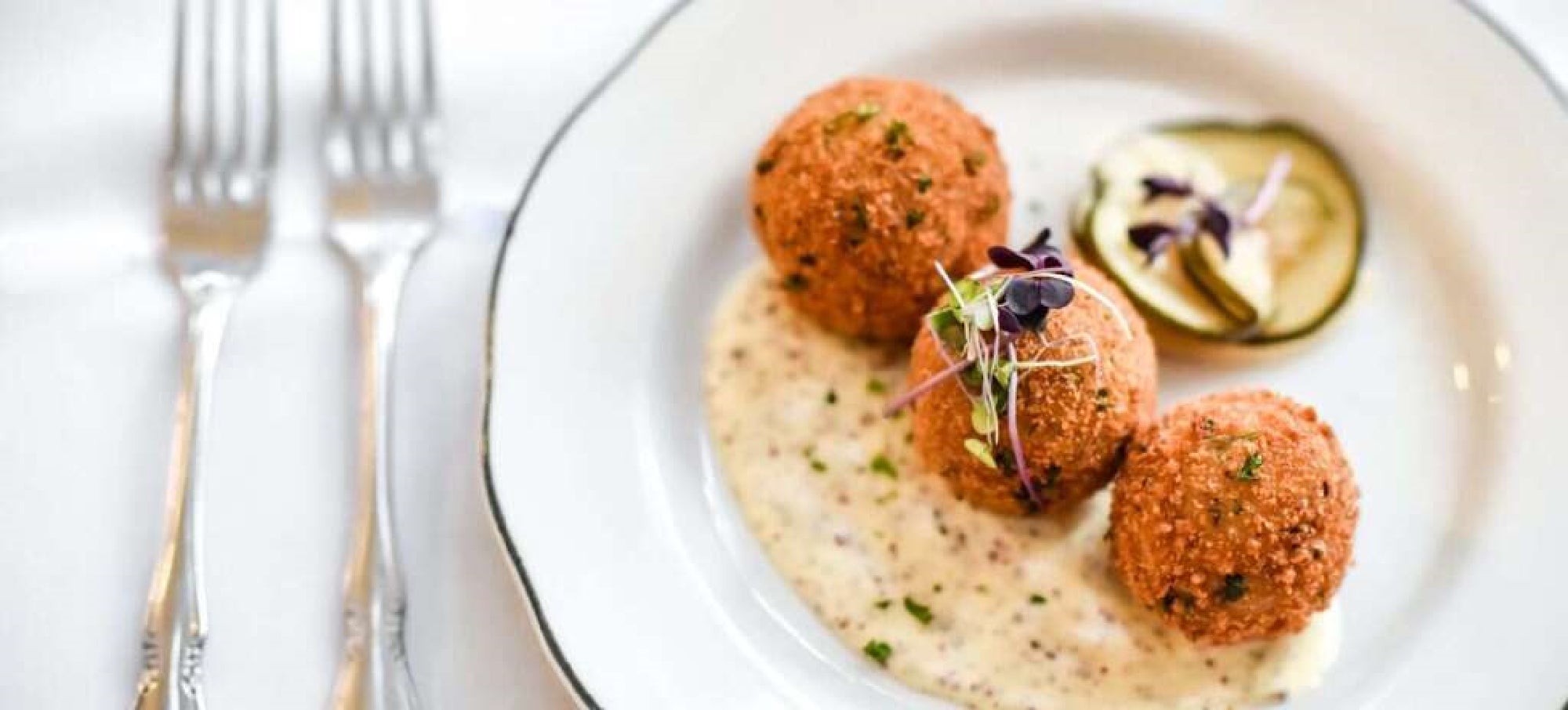 Three breaded and fried balls are served on a white plate with creamy sauce, garnished with microgreens and cucumber slices. Two forks are placed beside the plate.