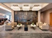 Modern hotel lobby with beige armchairs, marble tables, a black bench, and a wall-mounted fireplace set in a stone accent wall.