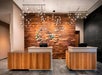 Modern hotel reception area with two wooden front desks, a wood-paneled wall featuring decorative stones, and ceiling lights arranged in a geometric pattern.