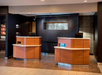Hotel reception area with two wooden counters, each equipped with a computer. Behind the counters is a dark wall with a decorative piece. To the left, shelves display various snacks.