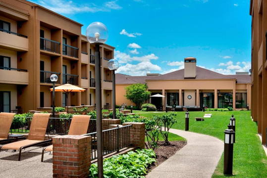 Courtyard of a hotel with green lawn, lounge chairs, and patio umbrellas, surrounded by multi-story buildings under a blue sky.