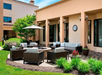 Outdoor patio with wicker furniture, a round fire pit, and umbrella, surrounded by plants and adjacent to a building with large windows and a wall clock.