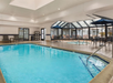 Indoor swimming pool area with a hot tub, surrounded by lounge chairs and tables with umbrellas. Skylights provide natural light.