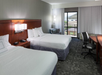 Hotel room with two double beds, a wooden headboard, desk with chair, floor lamp, and a window with curtains, offering an outdoor view.