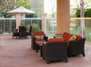Outdoor patio with wicker furniture featuring red cushions, a table, and an umbrella. The area is surrounded by a white fence and greenery.