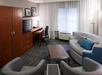 Hotel room with a work desk and TV, surrounded by a sofa and two chairs. Curtains cover the window, and two lamps provide lighting. The decor is modern with neutral tones.