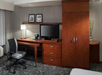 Hotel room workspace with a wooden desk, office chair, flat-screen TV, lamp, and framed photos on the wall. A matching wooden cabinet is on the right.