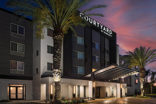 A modern hotel exterior with the "Courtyard by Marriott" logo, featuring palm trees and a sunset sky.