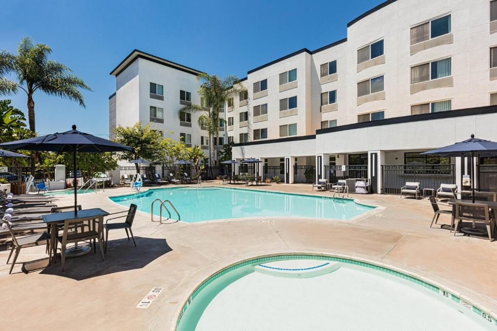 Outdoor hotel pool area with lounge chairs, umbrellas, and a hot tub, surrounded by a multi-story white building and palm trees under a clear blue sky.
