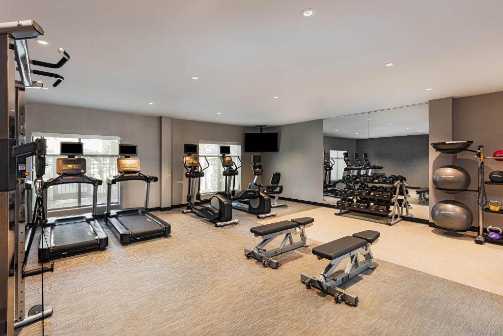 A fitness room with treadmills, exercise bikes, weight benches, dumbbells, and medicine balls. A large mirror and a wall-mounted TV are present.