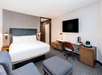 A modern hotel room with a king-sized bed, desk, chair, and wall-mounted TV. Room includes bedside lamps and a coffee maker on a cabinet. Flooring is carpeted in a neutral tone.