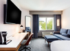 Hotel room with a bed, desk, chair, wall-mounted TV, and sofa. Large window with a view of palm trees. Coffee maker and lamp on the desk. Neutral color scheme.