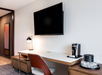 Modern hotel room desk setup with a large wall-mounted TV, a sleek lamp, a coffee maker, and a set of glasses.