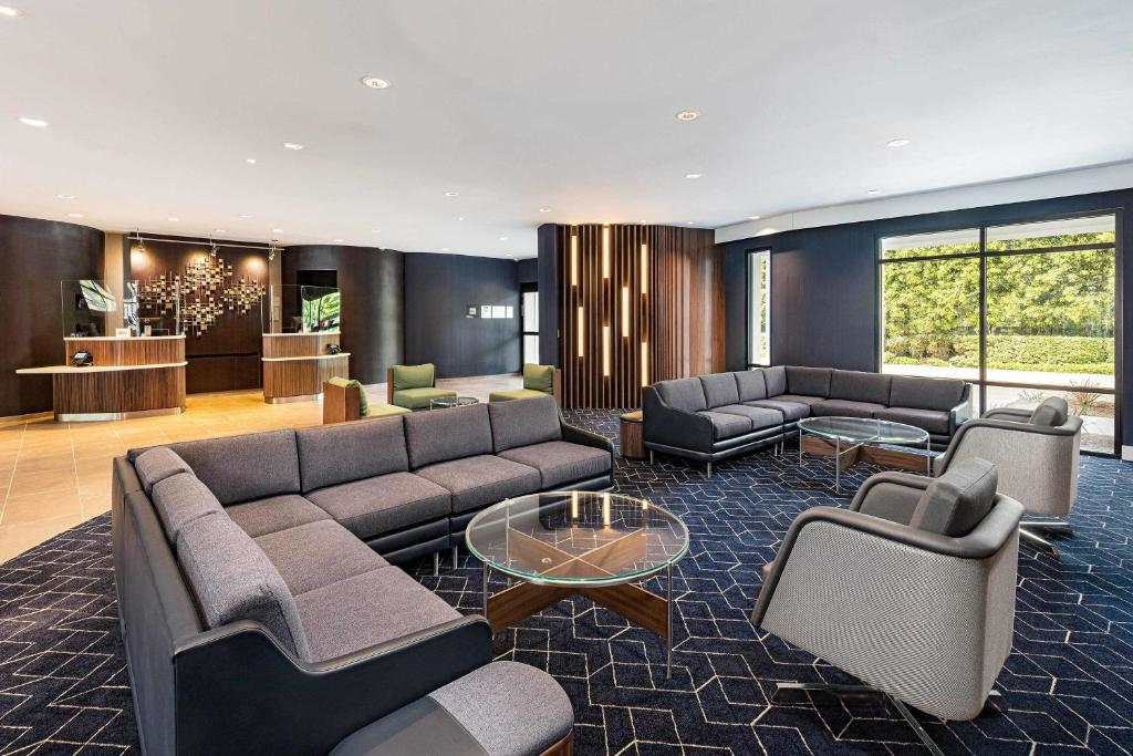 Modern hotel lobby with gray sofas, round glass tables, and a check-in counter in the background. Large windows provide natural light.
