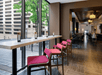 Modern cafe interior with a high table and pink stools near large windows. The view includes a geometric building and green trees. Warm lighting and wood flooring are present.