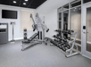 A gym room with a weight bench, dumbbell rack, exercise machine, medicine balls, a TV, and a scale. Walls are white, flooring is gray, and glass doors are on the right.