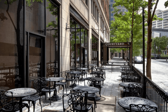 Outdoor seating area with empty wrought iron tables and chairs alongside a building. Trees line the sidewalk and a sign reads "Courtyard" near the entrance.