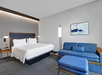Hotel room with a king-size bed, blue sofa, ottoman, two side tables with lamps, a large floor lamp, and a wall-mounted abstract painting.