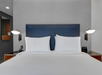 Modern hotel room with a large bed, blue headboard, white bedding, and symmetrical lamps on each side. A phone and a digital clock are on the wooden side tables.