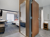 Hotel room with two beds, a work desk, and a visible bathroom with a walk-in shower. Neutral tones dominate the decor.