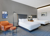Hotel room with two double beds, a gray wall, a floor lamp, an orange chair with a footstool, and framed artwork. Carpeted floor and modern decor.