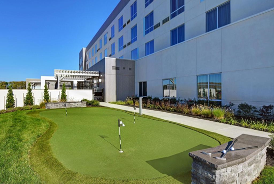 Hotel exterior with a small putting green, surrounded by a walkway, shrubs, and seating area.