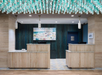 Hotel reception area with two wooden desks, modern light fixtures, and an abstract painting on a blue wall. Decorative elements hang from the ceiling above.