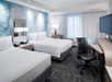 Hotel room with two double beds, a desk with a chair, a TV, and a floor lamp by the window. The walls have abstract blue-gray designs.