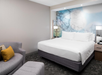 Modern hotel room with a king-sized bed, decorative accent wall, and grey upholstered furniture, including a chair and ottoman. Two bedside lamps illuminate the space.