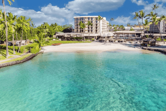 Beachfront resort with clear turquoise water, white sand, tall palm trees, and a multi-story hotel building under a partly cloudy sky.
