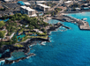 Aerial view of a coastal town with buildings near the shoreline, tropical greenery, a marina with docks, and clear blue ocean water.