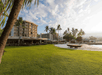 A beachfront hotel with palm trees, a vibrant green lawn, and a small boat on a sandy beach under a partly cloudy sky.