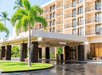 A multi-story hotel with balconies, a covered entrance, palm trees, and a paved driveway in front.