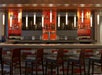 Empty bar with modern decor, featuring wooden stools, hanging lights, and shelves stocked with bottles and glasses behind the counter.