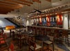 Bar interior with a long counter, stools, and tables with chairs. Modern lighting hangs from the ceiling. Stairs lead to an upper level, and a yellow couch is in the background.