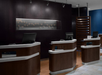 Hotel reception area with three modern, dark wooden desks equipped with computers, set against a dark wall with a decorative piece.